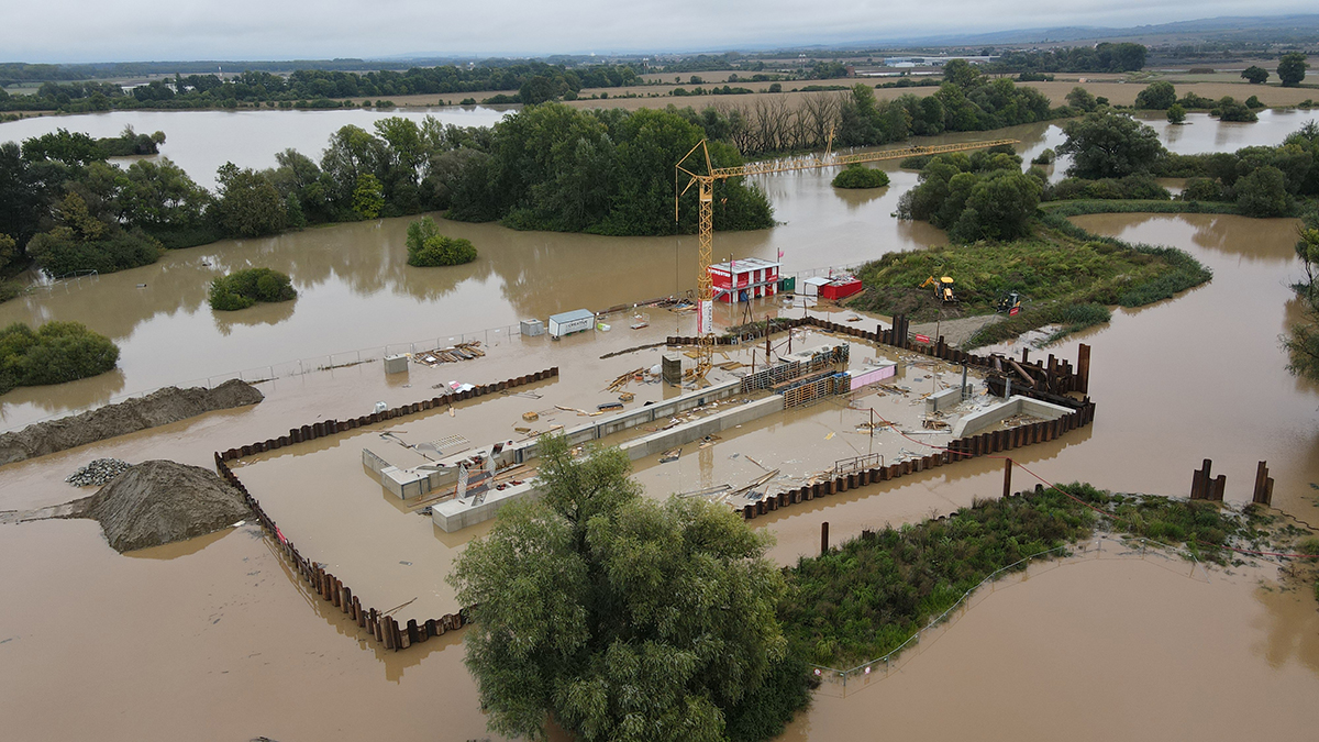 Stavební projekt na rozvodněné ploše, kde jsou stavební základy částečně ponořené pod hladinou. Pracovníci a technika jsou soustředěni kolem rozestavěné konstrukce, zatímco okolní příroda je pokryta rozlévající se vodou. Je to ukázka stavebních výzev v zátopových oblastech.