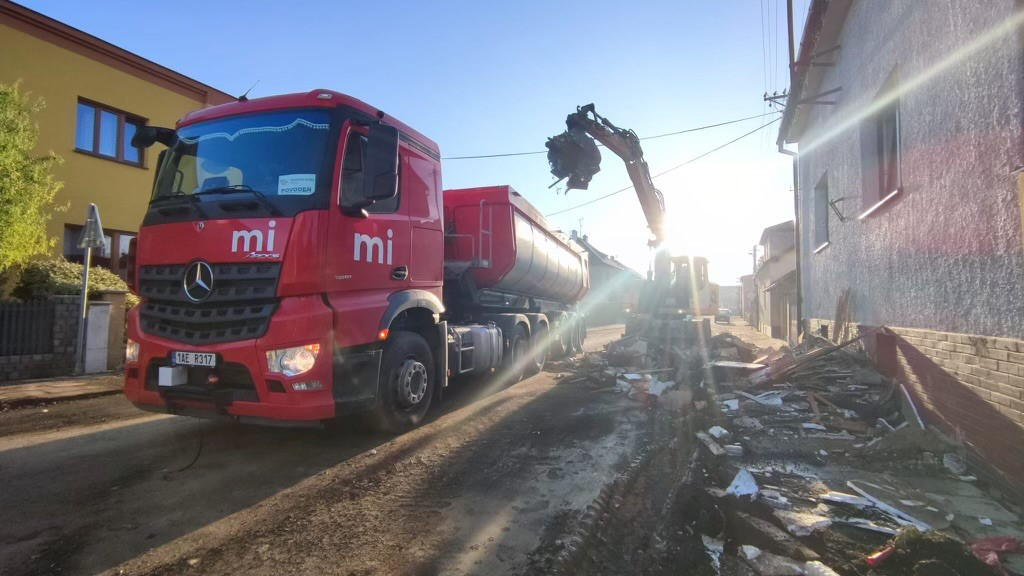 Červený nákladní vůz značky Mercedes parkuje vedle demolice v obci, kde probíhá úklid trosek. Bagr nakládá odpad do korby vozu, zatímco ranní slunce vrhá dlouhé stíny a osvěcuje celý pracovní prostor. Scéna působí klidně, přestože probíhá intenzivní stavební činnost.