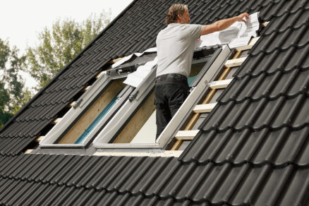 Montáž střešních oken VELUX na černé střešní krytině. Pracovník instaluje nové okno pro zvýšení energetické úspory.